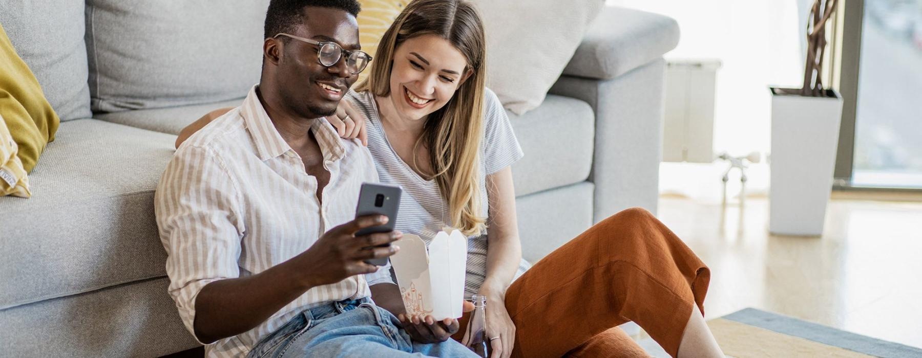 a man and woman with take out, sit against a couch on their living room floor and watch at their cell phone